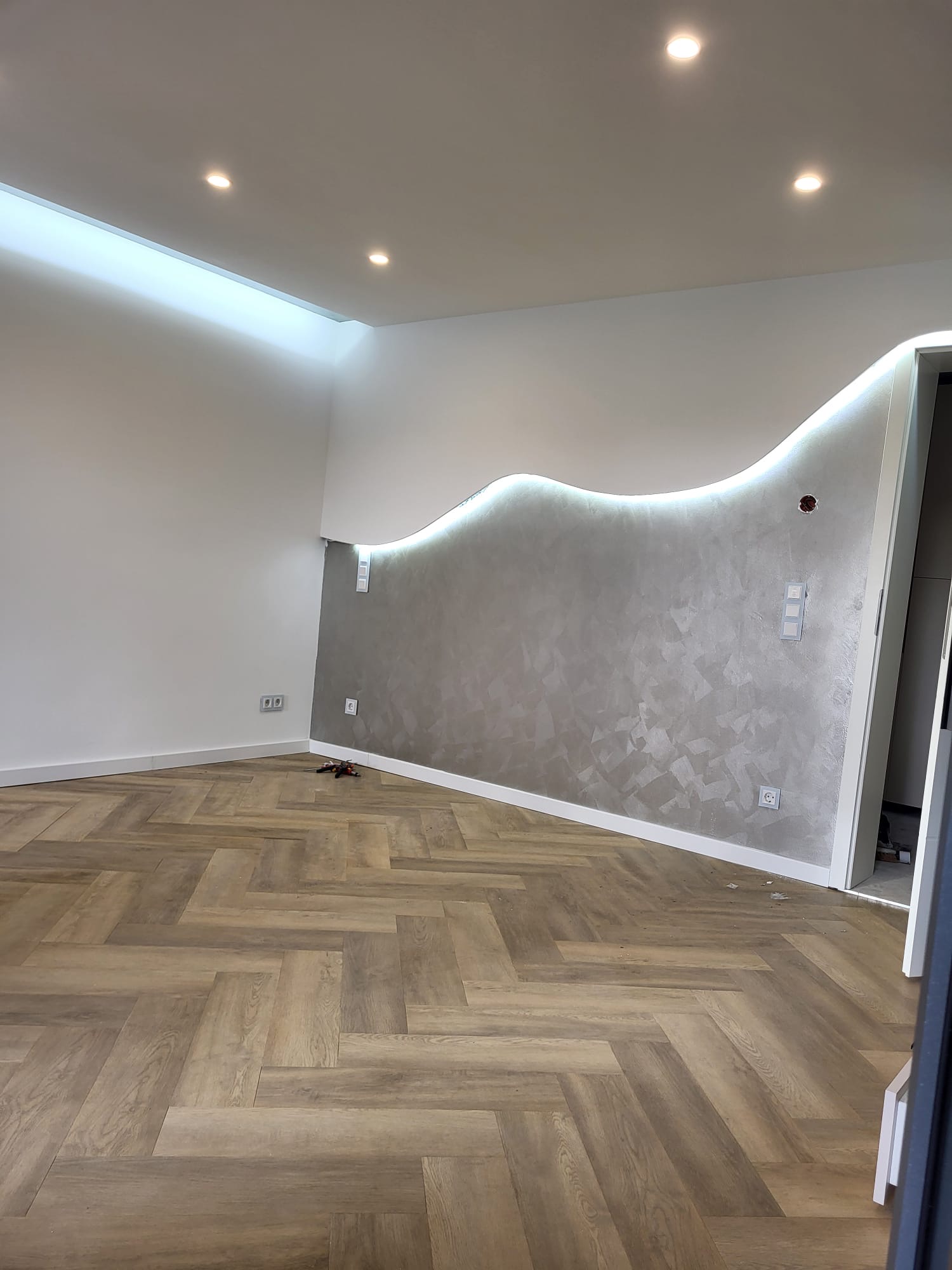 Wohnraum mit silberner Effektspachtel-Designwand, indirekter LED-Beleuchtung und Fischgrät-Vinylboden