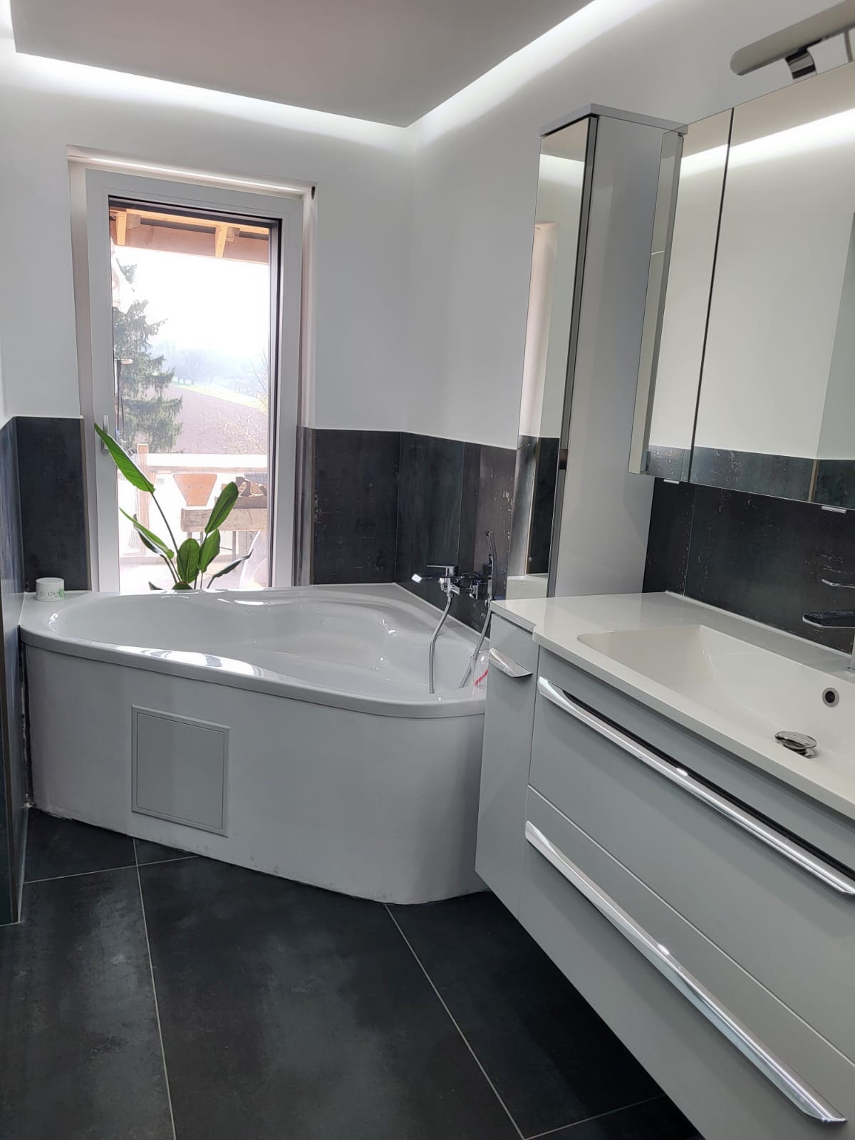 Saniertes Badezimmer mit weißer Eckbadewanne, anthrazitfarbenen Großformatfliesen und LED-Lichtvoute
